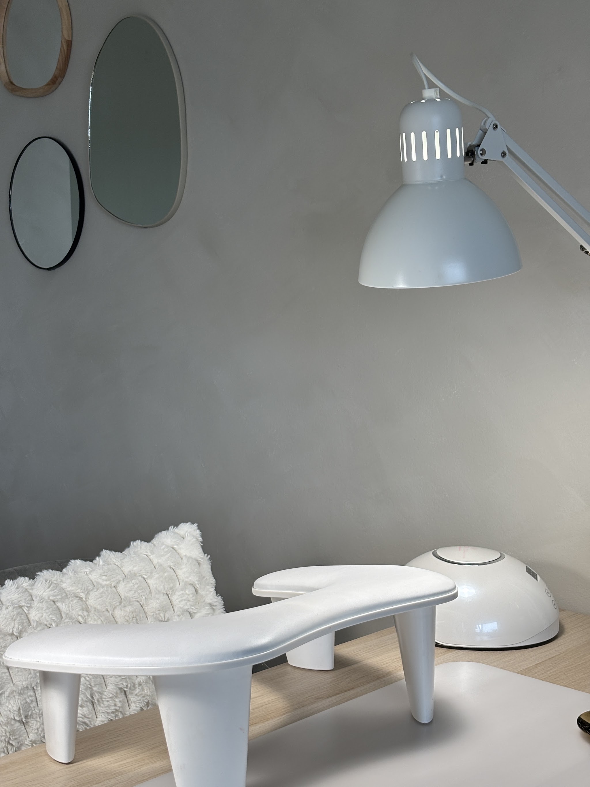 Werkplek met een witte nageltafel, een lamp en ronde spiegels tegen een betongrijze muur